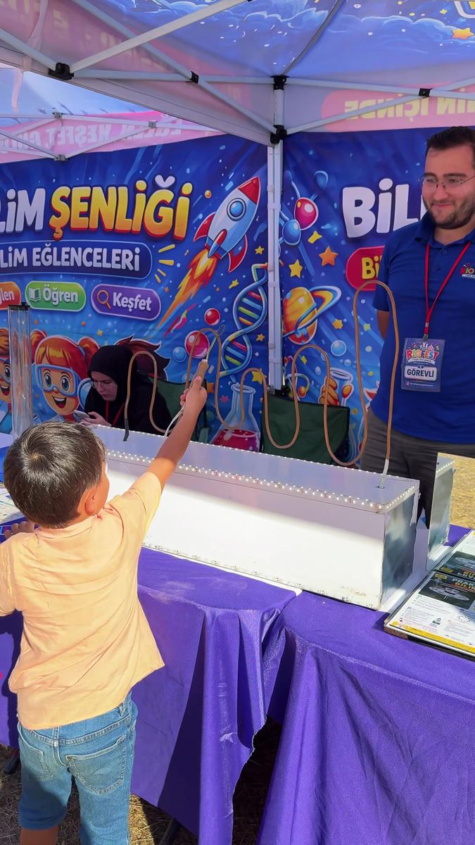 Bilim eğlenceleri standında bir çocuk, görevli eşliğinde bilim cihazını deniyor
